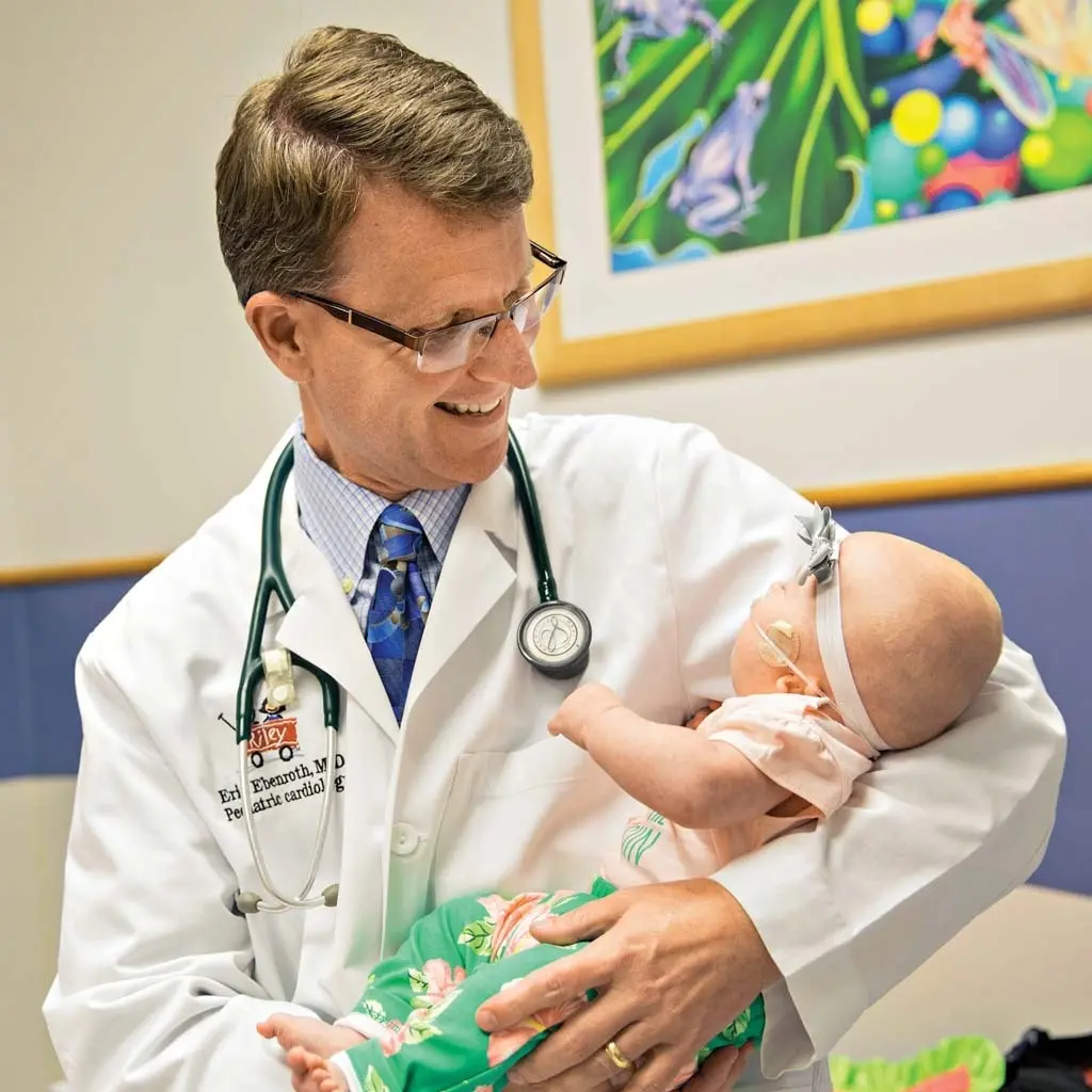 A male pediatrician in glasses and a lab coat, holding and smiling at a baby wrapped in a floral blanket in a clinic room with colorful wall art.