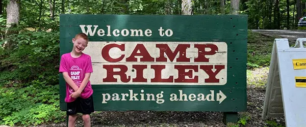 Welcome to Camp Riley Sign with Luke standing in front.